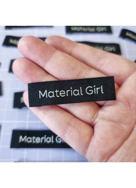 A hand holding a narrow, black sew-in label featuring the words 'Material Girl' in metallic silver