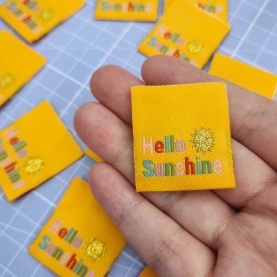 A hand holding a bright yellow sew-in label featuring a metallic gold sunshine and the words 'Hello Sunshine'
